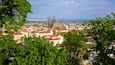 A cityscape with prominent twin spires of a cathedral, surrounded by lush green trees and various buildings with red roofs.