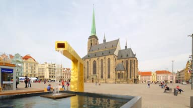 Uma igreja histórica com uma torre alta, uma fonte com braço dourado e uma praça movimentada com pessoas e edifícios.