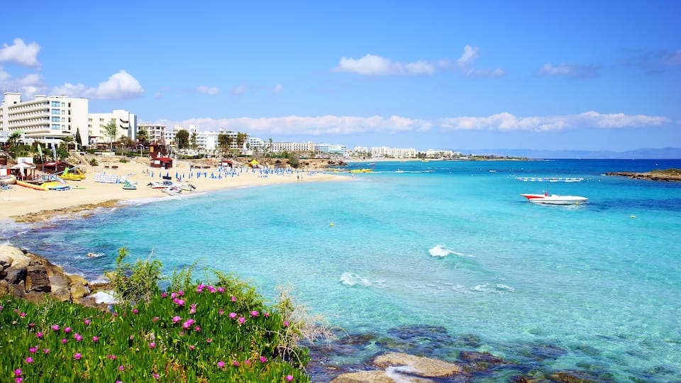 A coastal beach with clear turquoise waters, a rocky shoreline, and a row of buildings in the background.