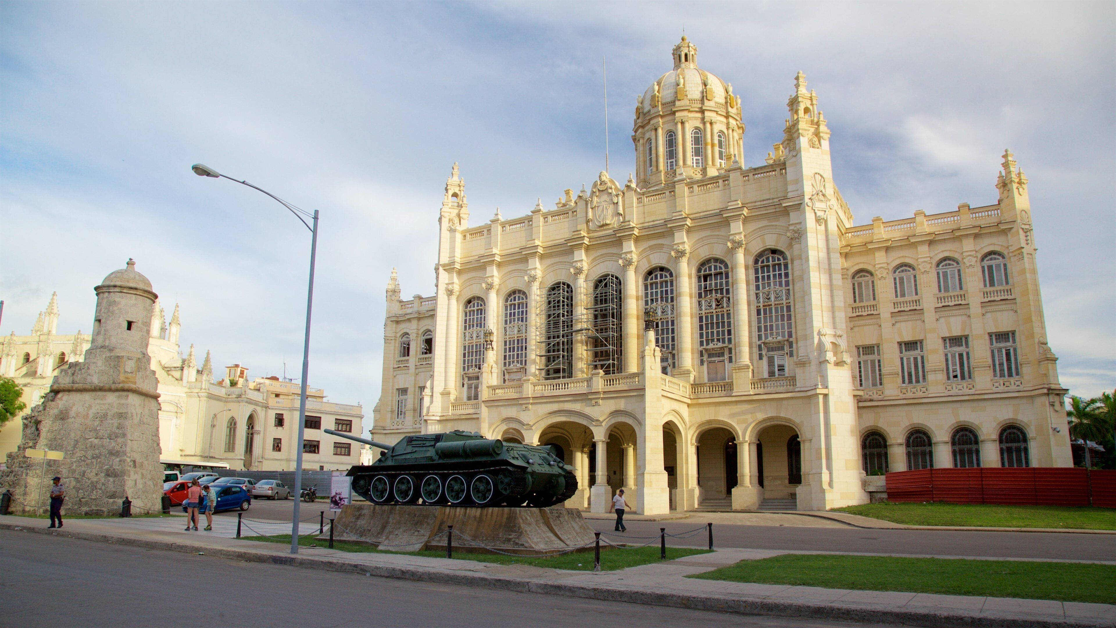 Un edificio histórico con arquitectura ornamentada, un gran monumento en forma de tanque al frente y un cielo despejado.