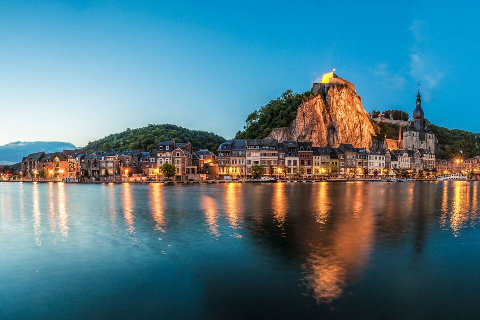 Une rivière paisible où sont amarrés des bateaux, une montagne imposante surmontée d’une église, et une ville bordée de bâtiments historiques le long de la rive.