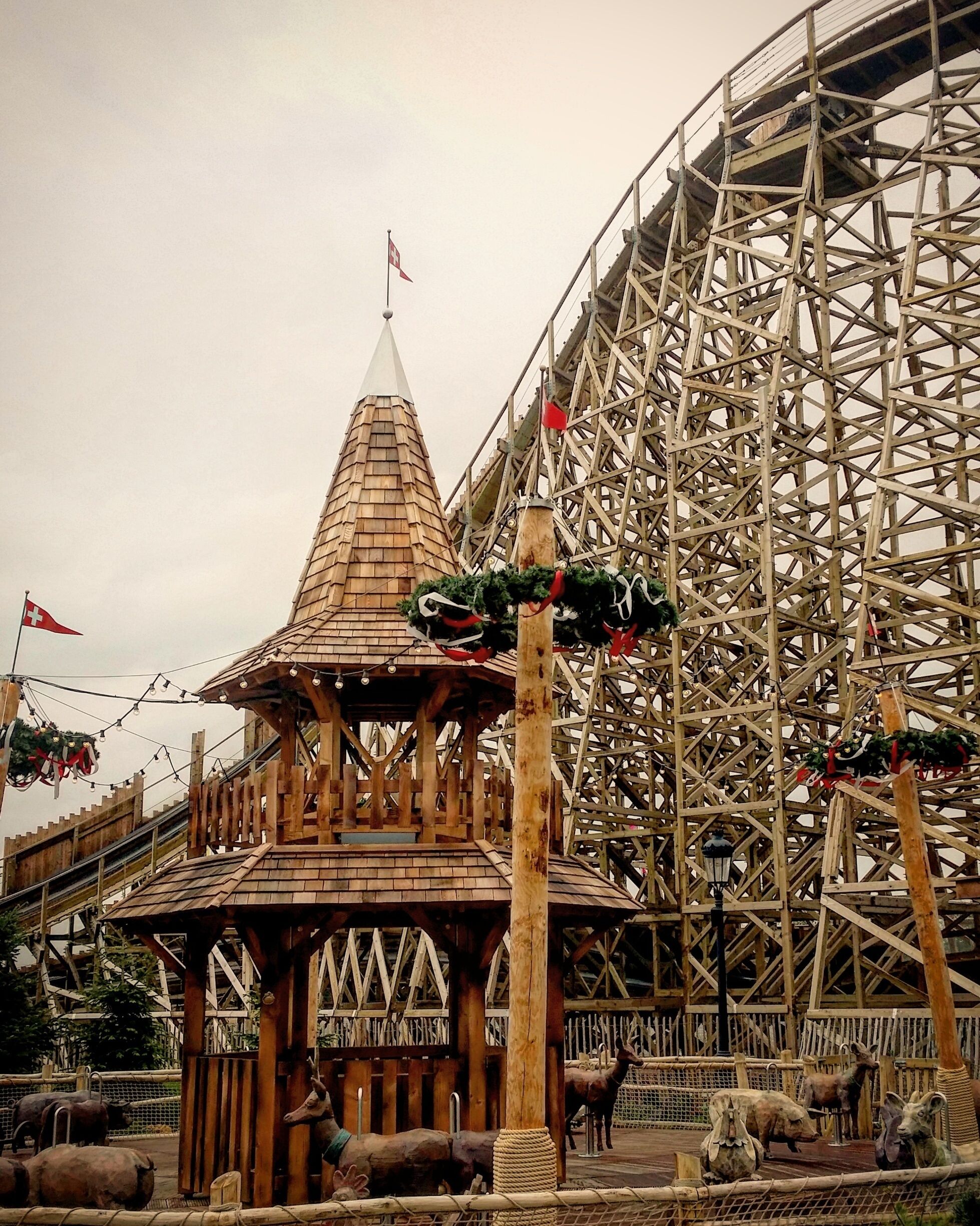 A wooden structure with a conical roof, surrounded by a wooden fence, and a large wooden roller coaster in the background.