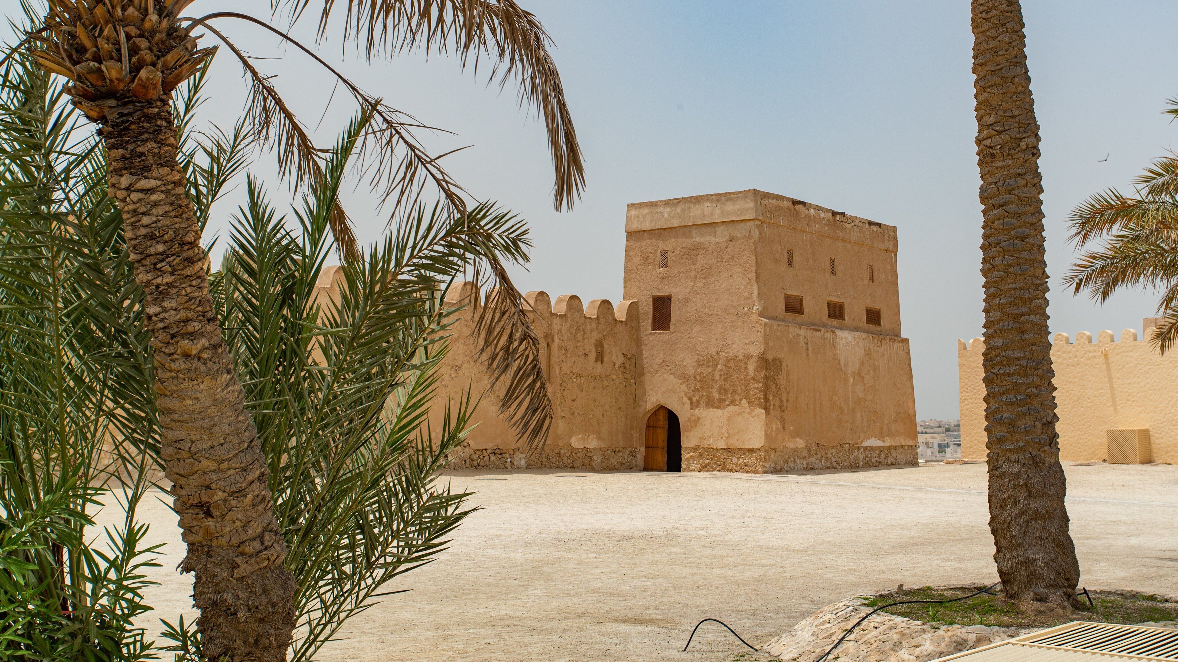 A historic building with a large arched entrance, surrounded by palm trees and sandy ground.
