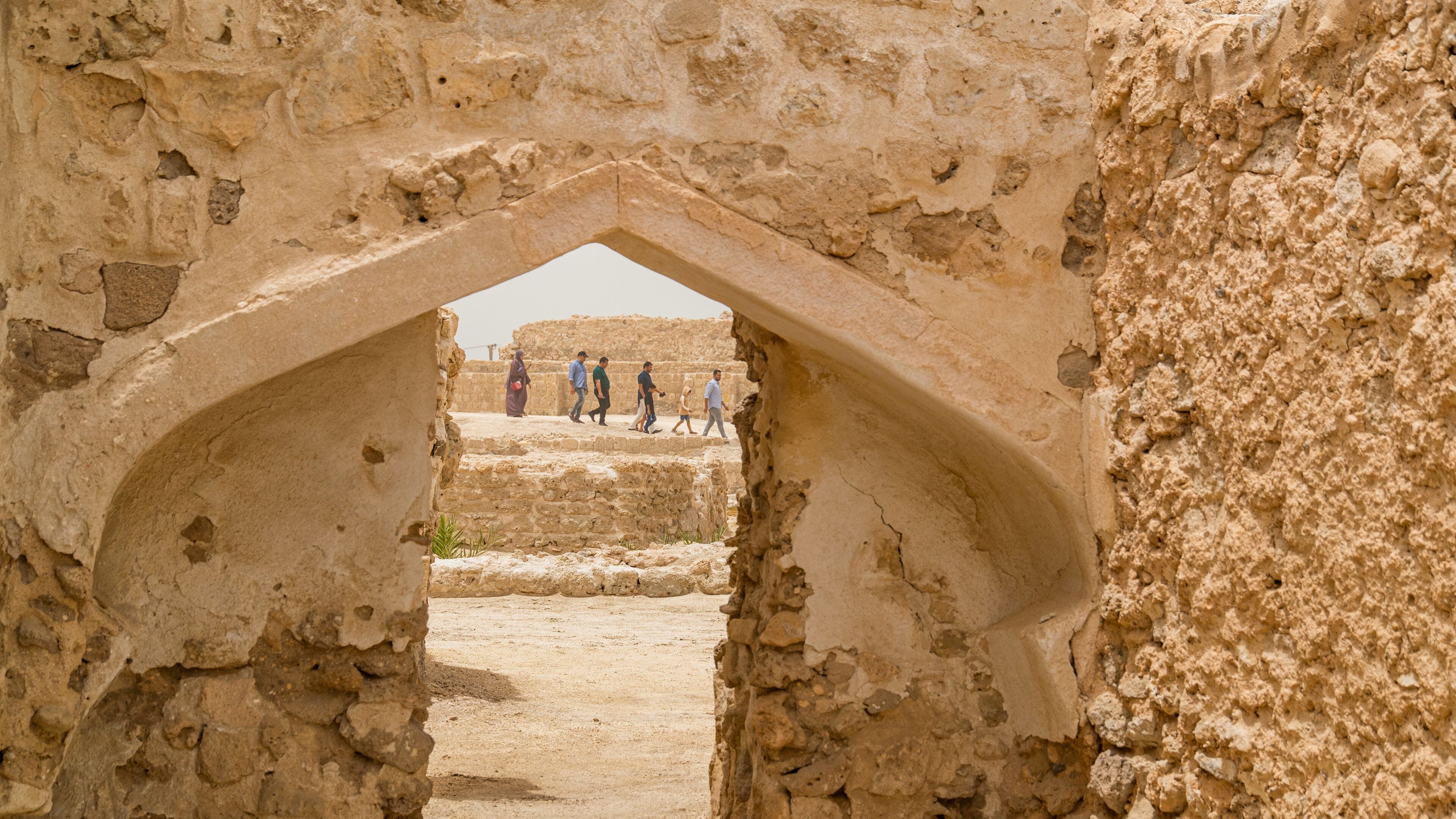 An ancient stone archway leading to a historical site with ruins and people walking around.