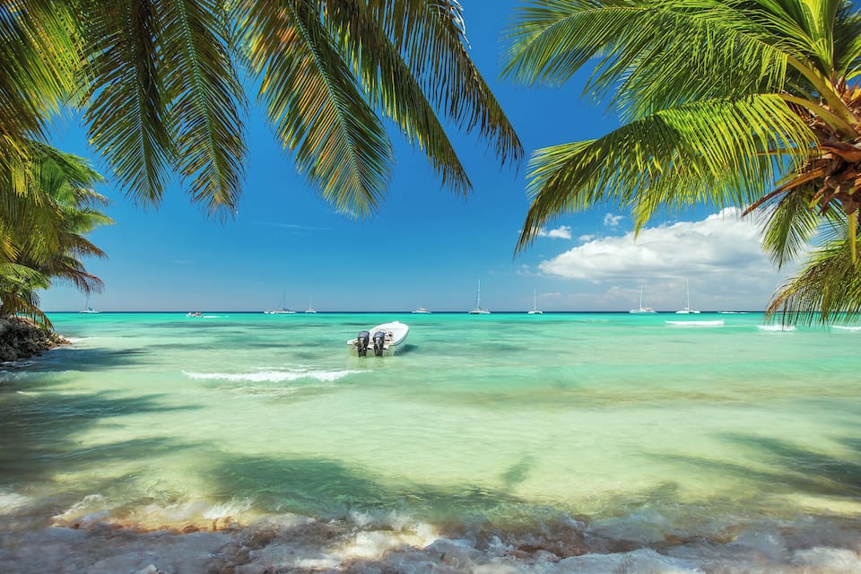 Helder turquoise water met een boot, palmbomen en zeilboten in de verte.