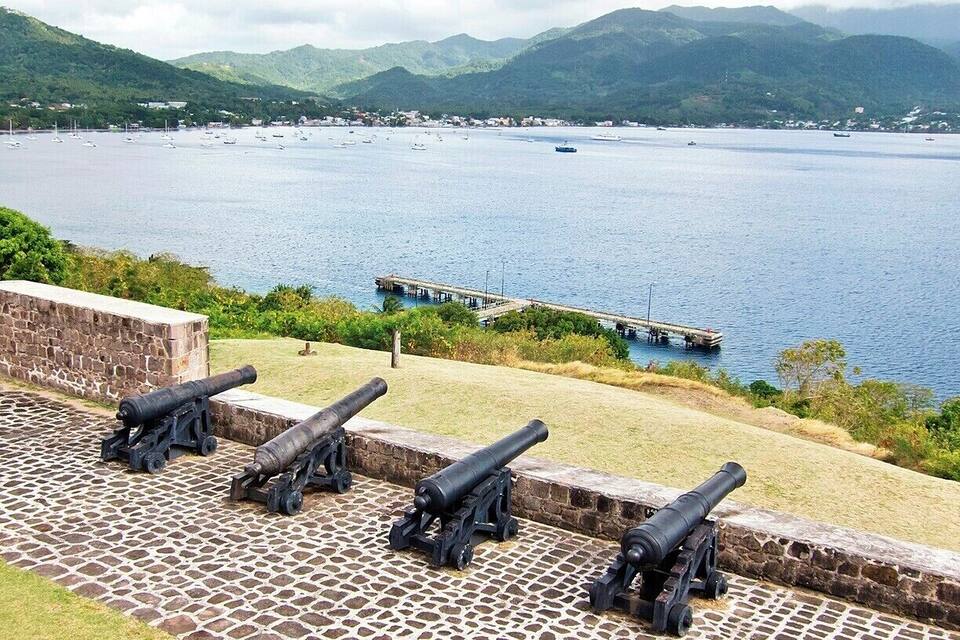 A coastal landscape with a stone wall and cannons overlooking a bay with boats and a pier.