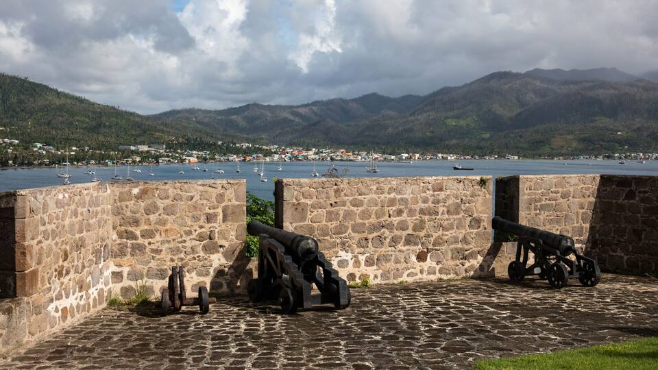 A coastal fort with cannons overlooking a harbor with boats and a mountainous backdrop.