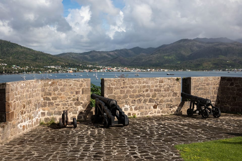 A coastal fort with cannons overlooking a harbor with boats and a mountainous backdrop.