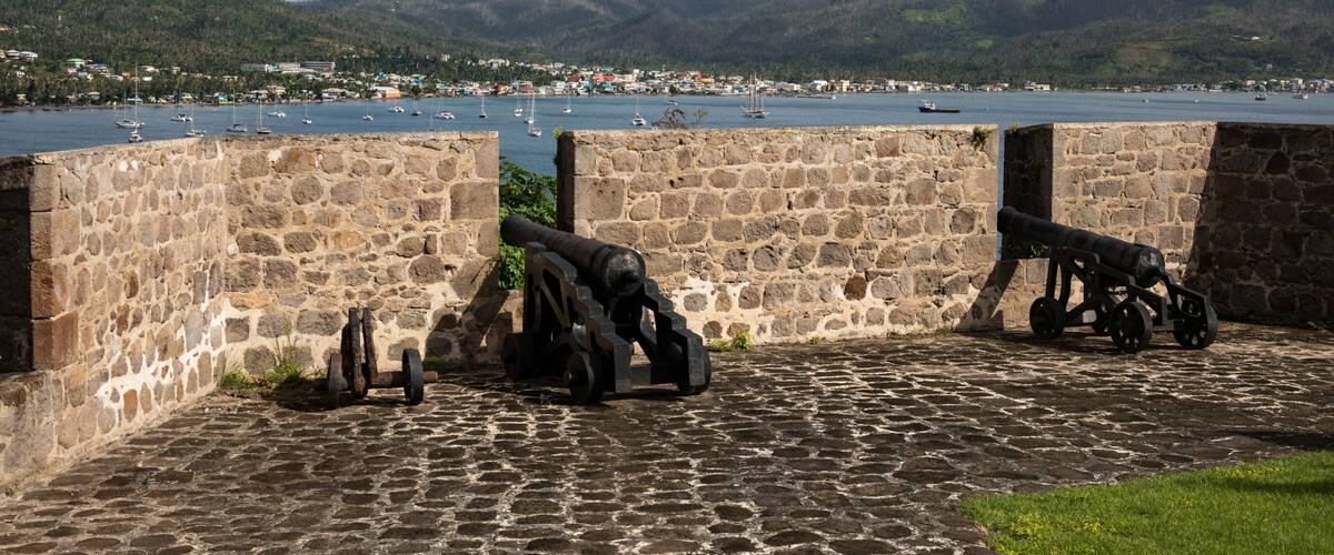 A coastal fort with cannons overlooking a harbor with boats and a mountainous backdrop.