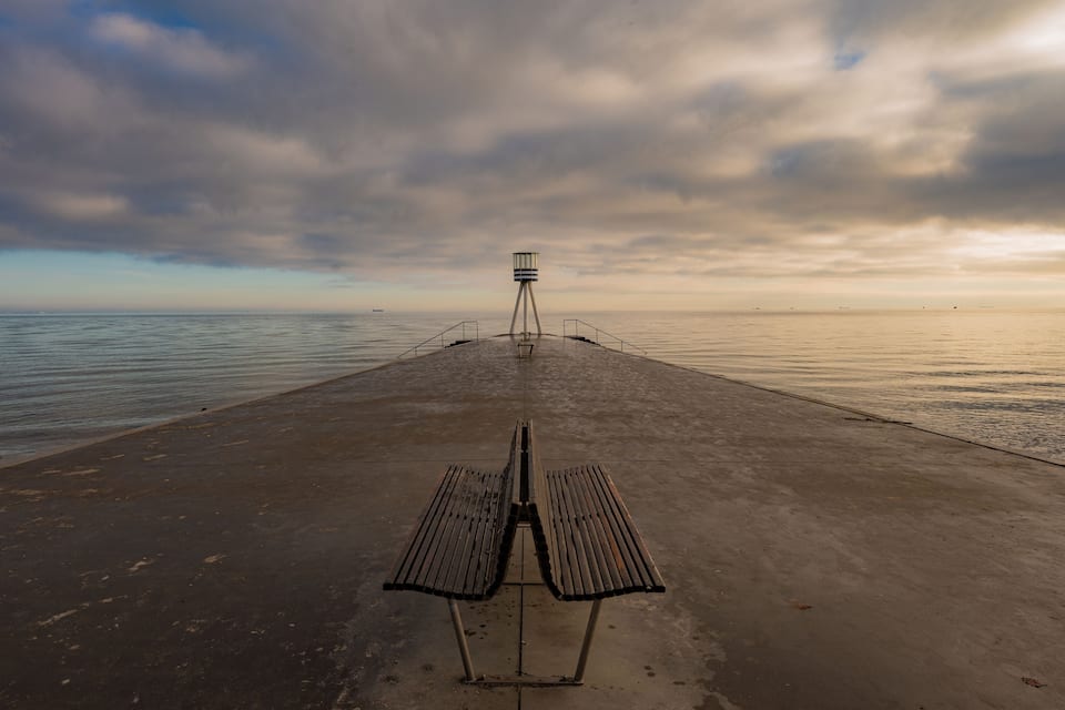 A concrete pier with a bench and a lighthouse in the distance.