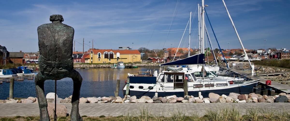 A statue of a man in a chair by a calm harbor with boats and buildings in the background.