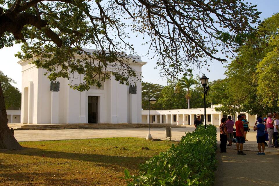 A white building with columns, surrounded by greenery and a group of people standing nearby.