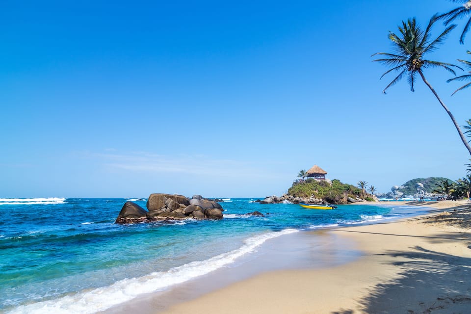 Una playa con aguas azul cristalino, una cabaña con techo de paja y palmeras.