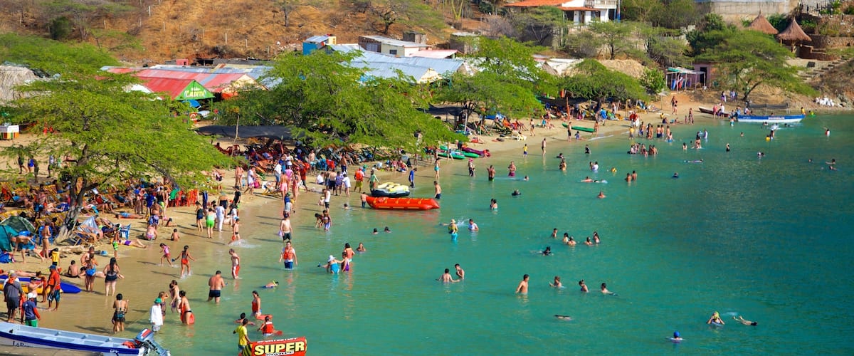 A crowded beach with people swimming and sunbathing, surrounded by houses and trees.