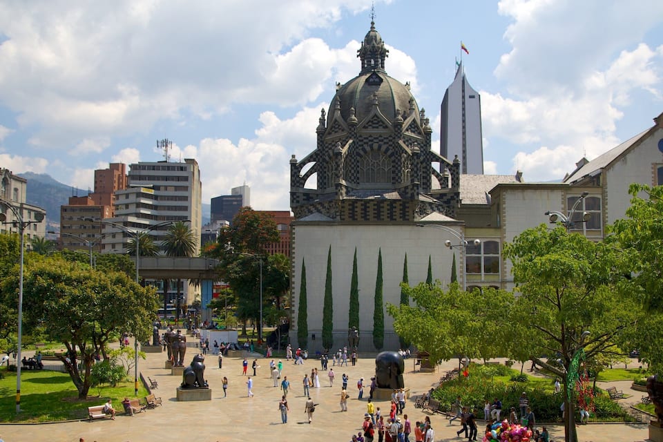 Uma praça com um edifício grande, com cúpula e torres, rodeada por árvores e pessoas.