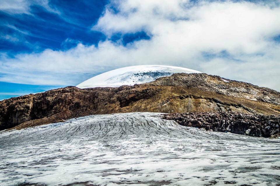 A snow-covered mountain with a glacier in the foreground and a clear blue sky with clouds.