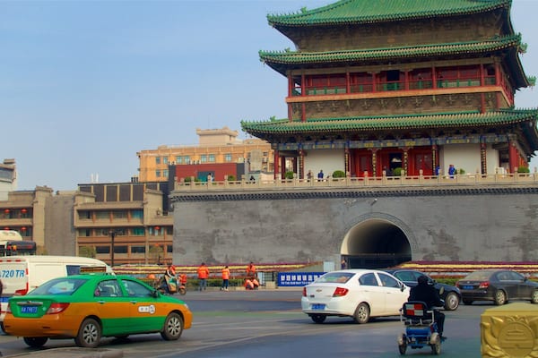 A traditional Chinese gate with a large archway, a green-roofed building, and modern vehicles on the road.