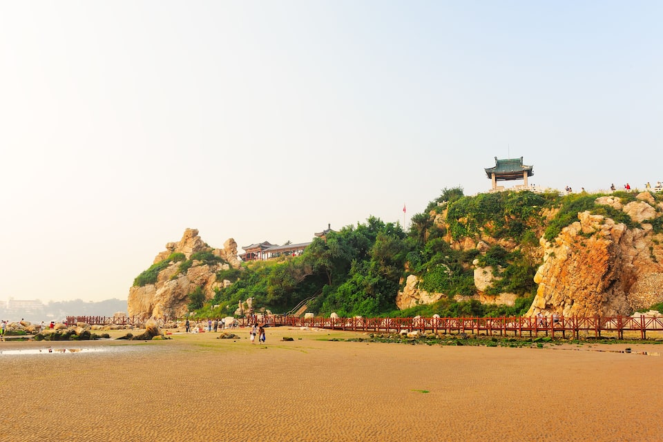 A beach with a traditional pavilion on a rocky hill, a wooden fence, and people walking.