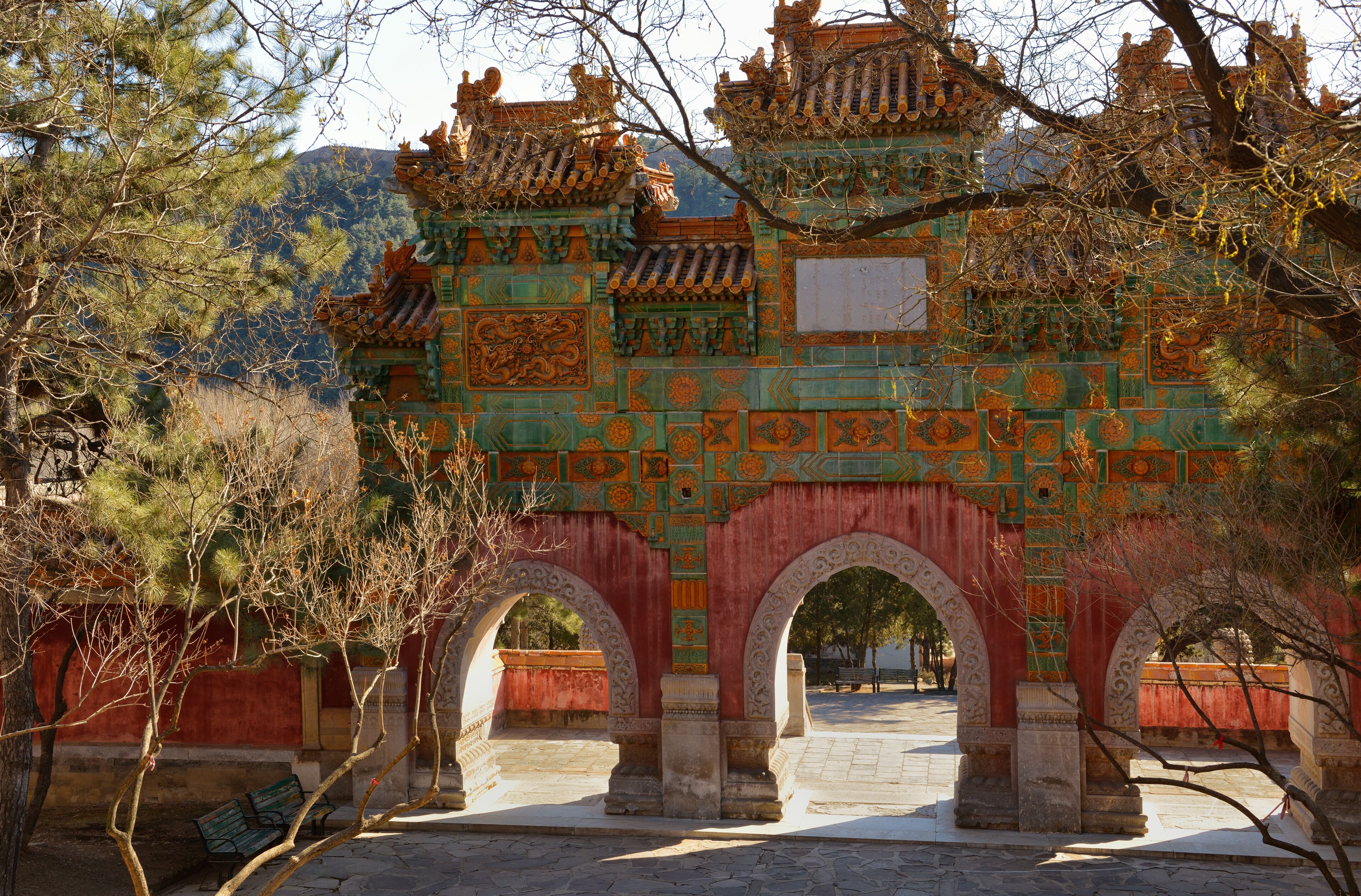A traditional Chinese gate with ornate designs and a red background, surrounded by trees and a mountainous landscape.