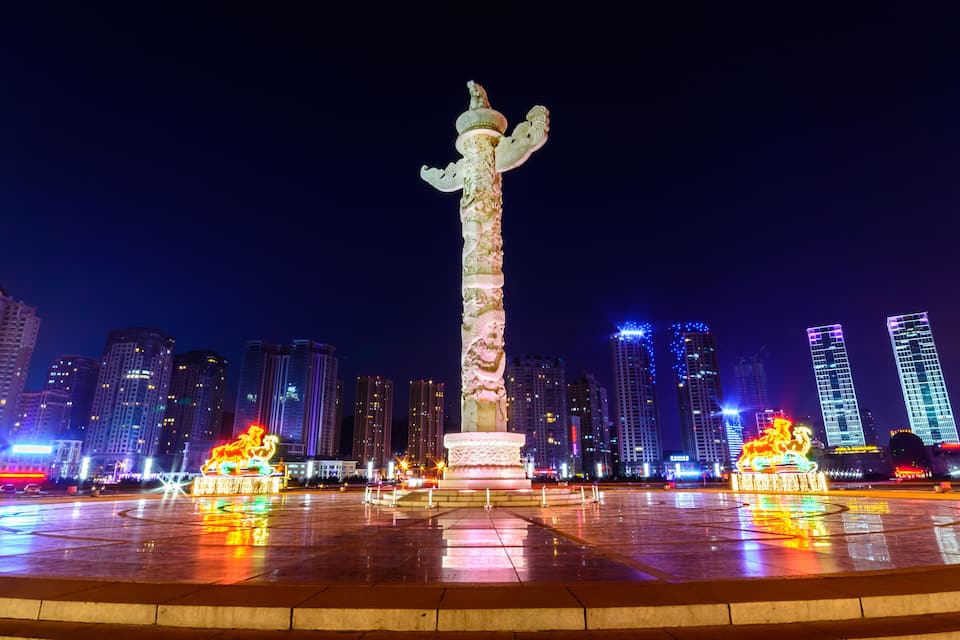 Paisagem urbana noturna, com um monumento iluminado em destaque no centro, rodeado por edifícios altos e modernos.