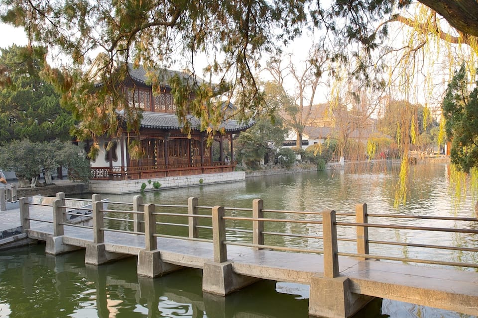 Um pavilhão tradicional chinês à beira de um lago tranquilo, com uma ponte de madeira e árvores exuberantes.