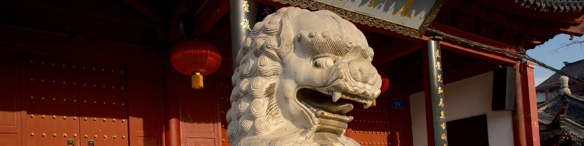 A stone lion statue at the entrance of a traditional Chinese building with red doors and a tiled roof.