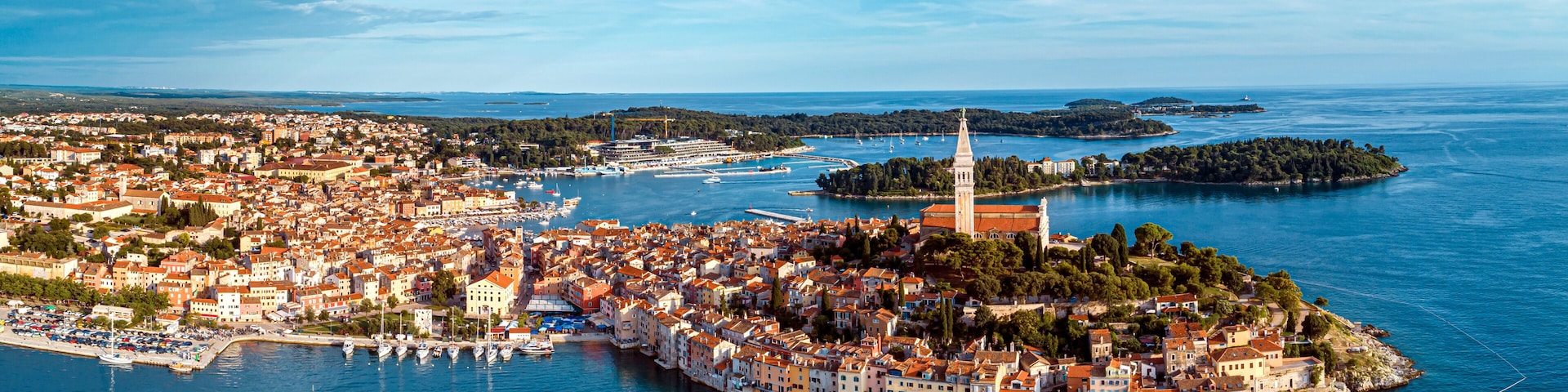 Una ciudad costera con una torre destacada, un puerto con barcos y una pequeña isla de vegetación densa.