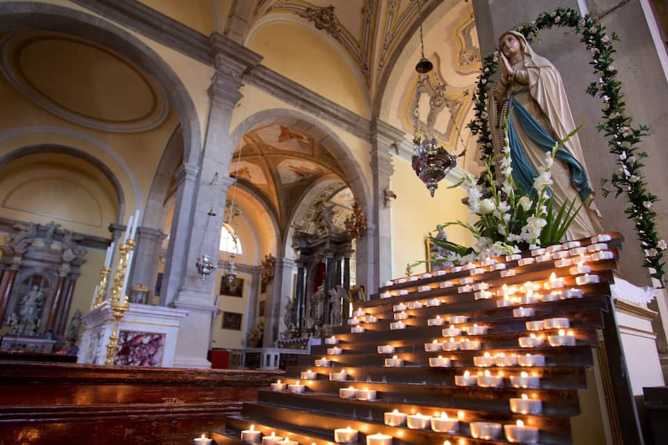 Interior de igreja com estátua, velas e arquitetura ornamentada.