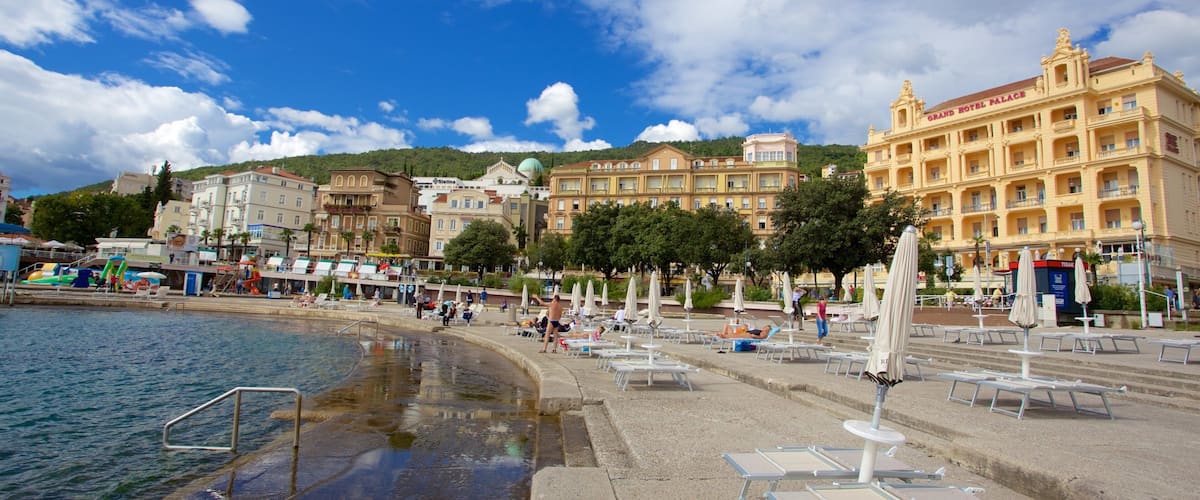A coastal promenade with beach chairs and umbrellas, a swimming pool, and historic buildings including the Grand Hotel Palace.