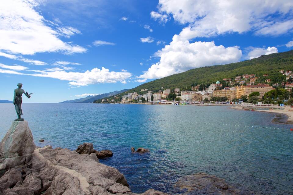 A coastal town with a statue on a rocky outcrop, clear blue sea, and a hillside with buildings.