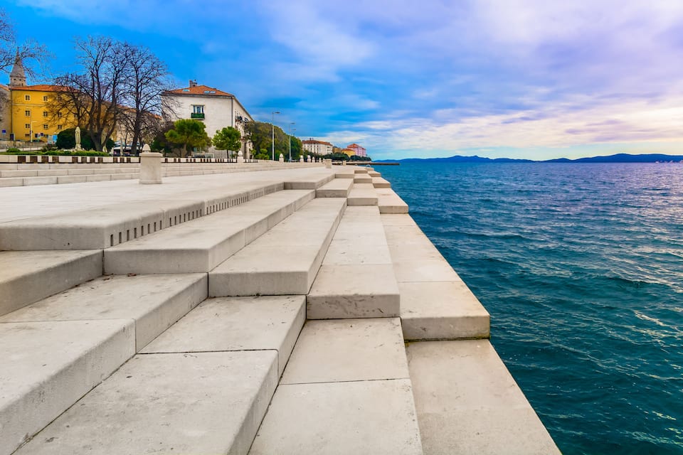 Um calçadão à beira-mar com escadas que descem até a água, edifícios e um céu claro.
