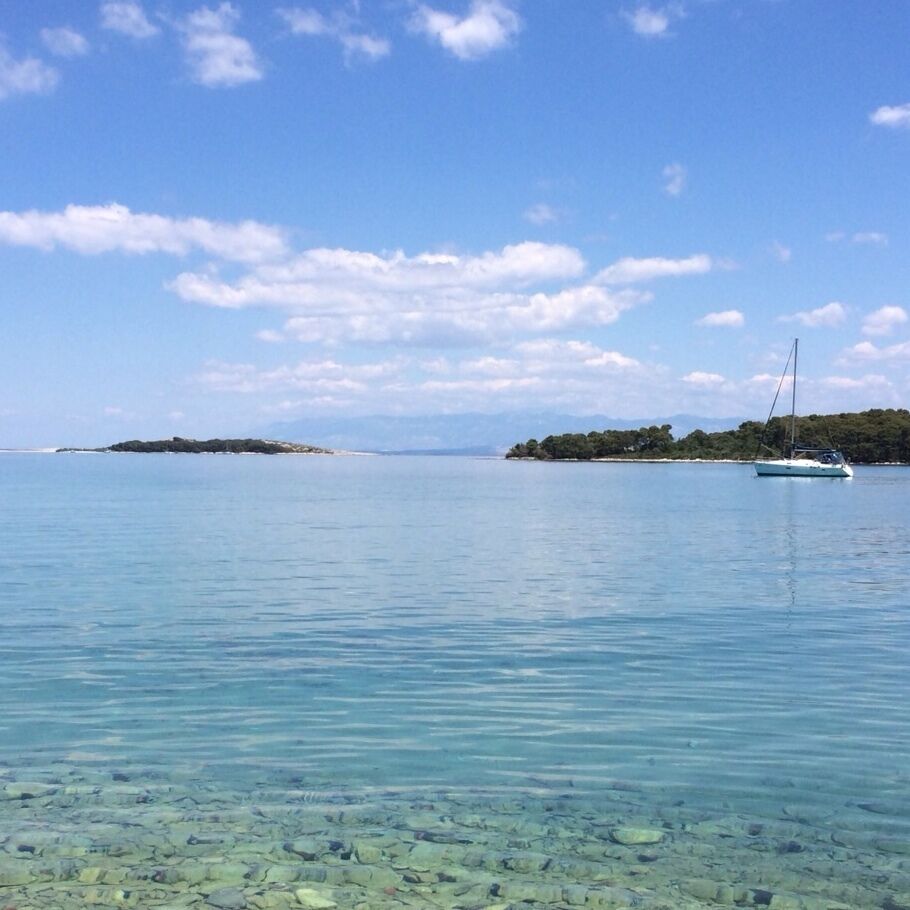 A calm sea with a sailboat, clear water revealing the seabed, and distant islands under a blue sky.
