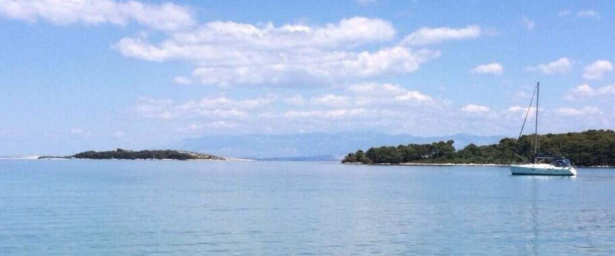 A calm sea with a sailboat, clear water revealing the seabed, and distant islands under a blue sky.
