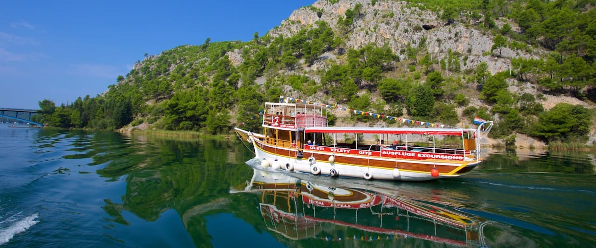 A boat with "IZLETI VLEYTY" and "AUSFLUGE EXCURSIONS" on its side, floating on calm water with a rocky hillside in the background.
