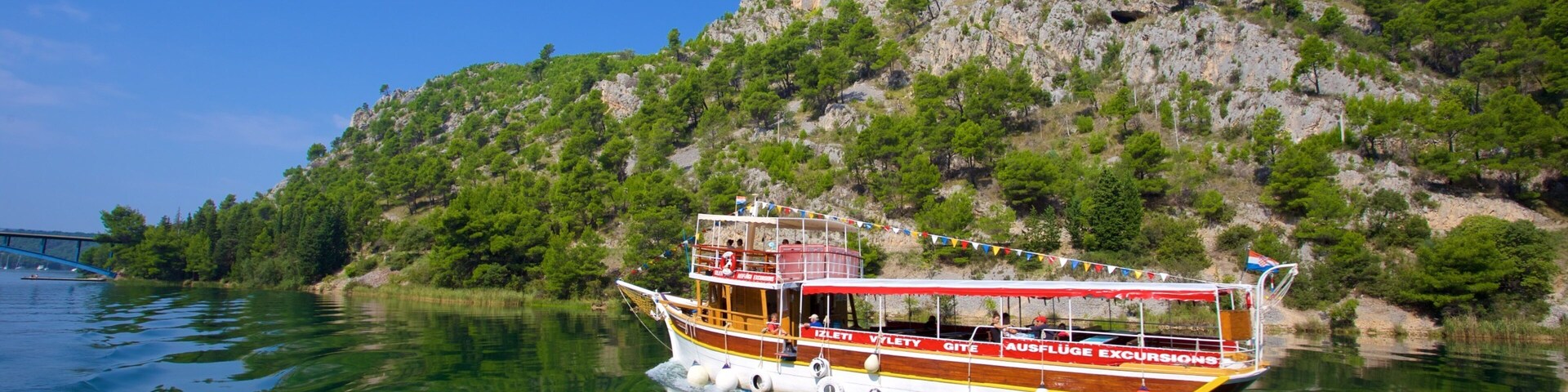 A boat with "IZLETI VLEYTY" and "AUSFLUGE EXCURSIONS" on its side, floating on calm water with a rocky hillside in the background.