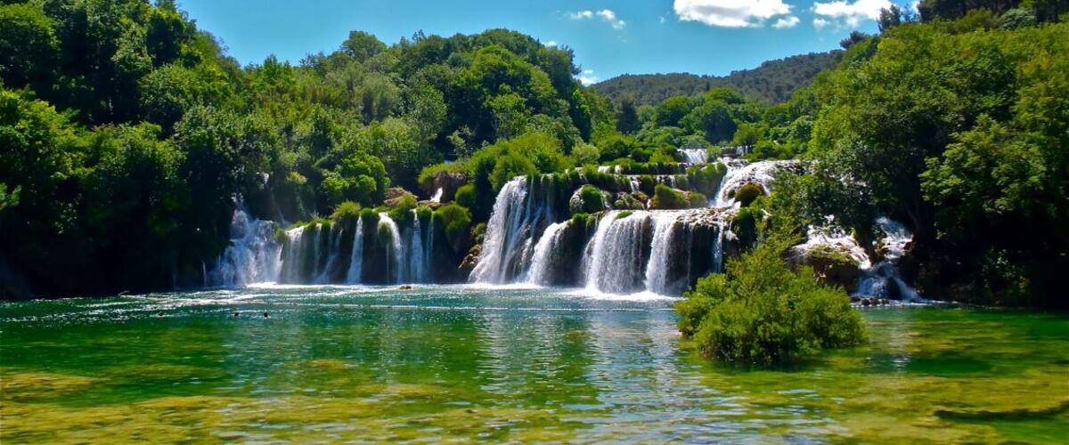 A multi-tiered waterfall surrounded by lush greenery and clear blue water.