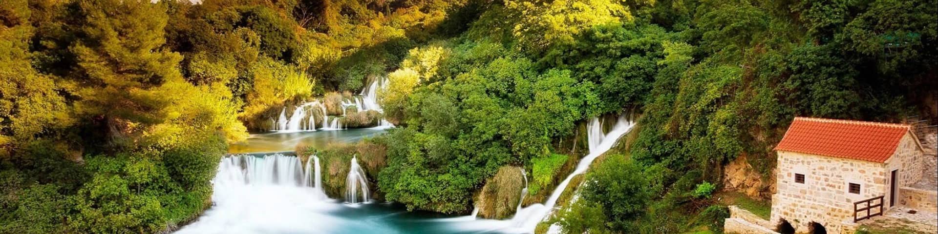 A river with waterfalls, surrounded by lush greenery and a stone building with a red roof.