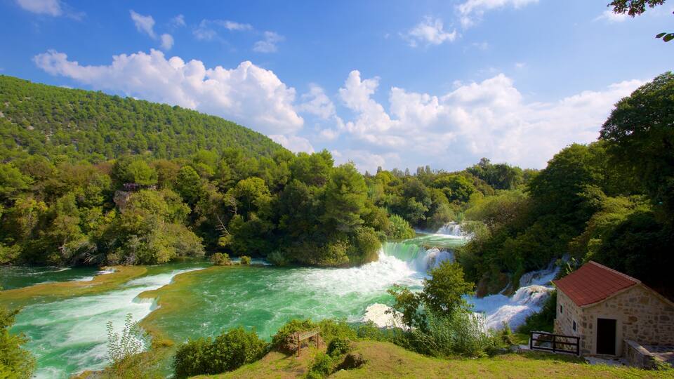 A river with waterfalls, surrounded by dense green forest and a stone building.