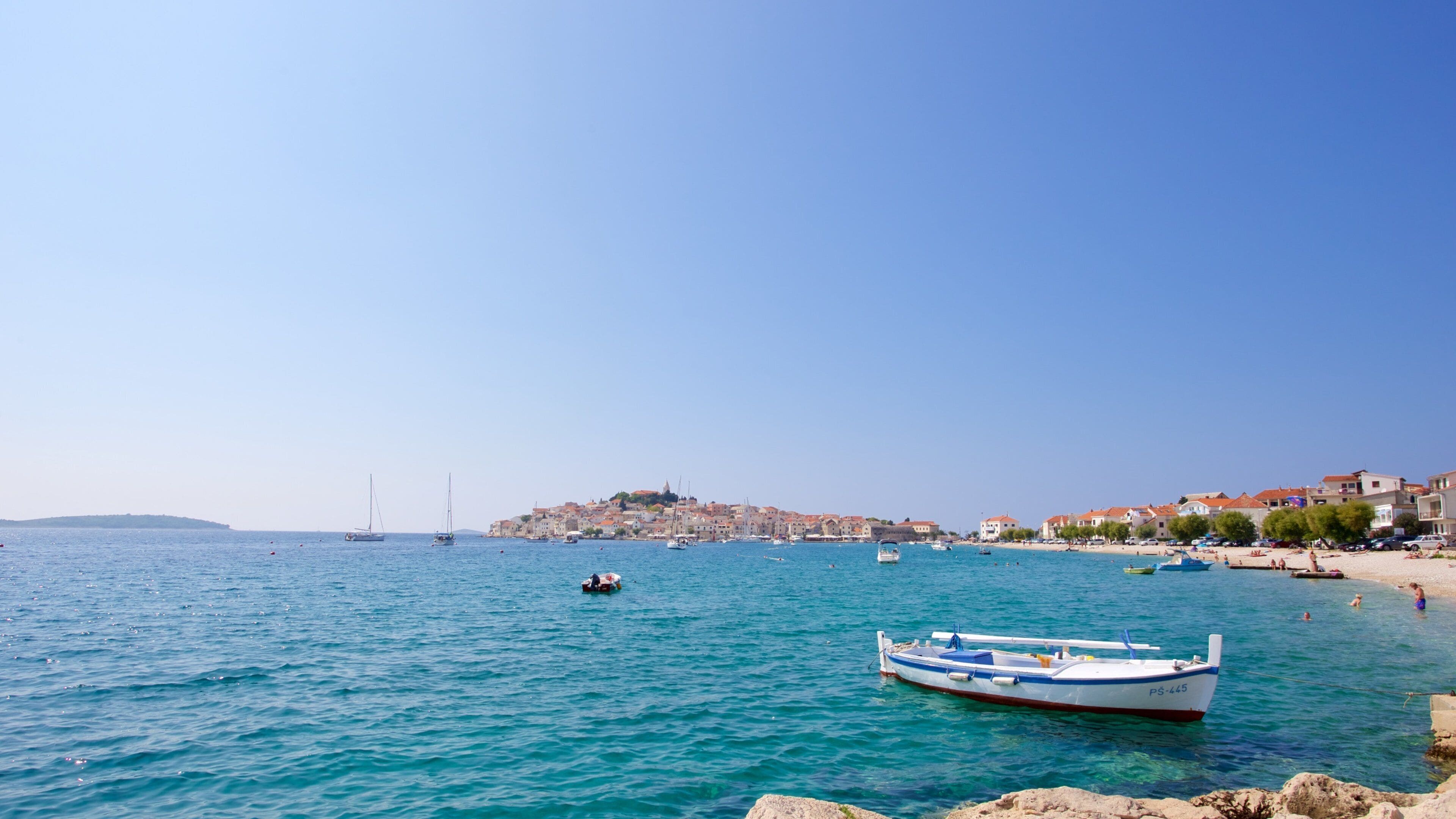 A coastal town with clear blue waters, a prominent white boat with blue stripes, and a distant hilltop with buildings.