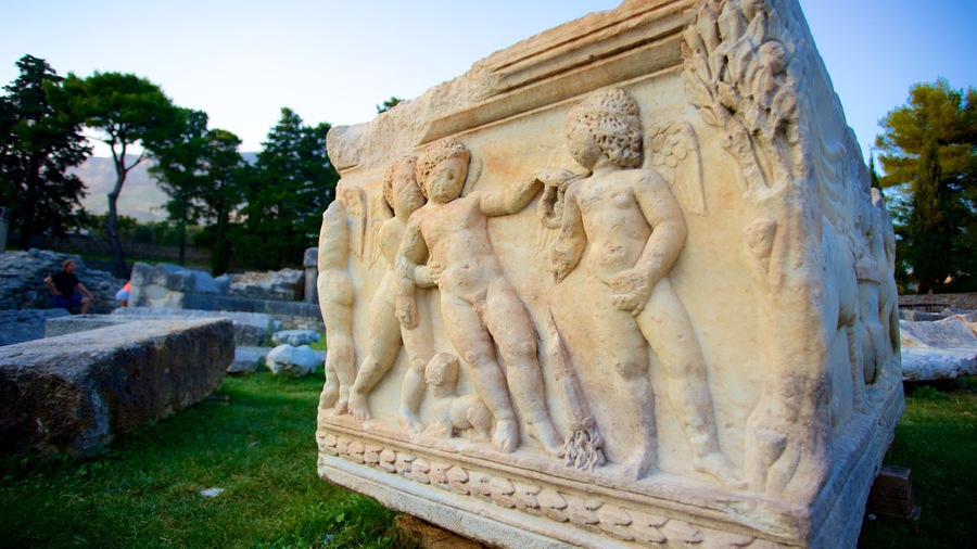 Una escultura de mármol con tres figuras en estilo clásico, ubicada en una zona verde con árboles y ruinas al fondo.