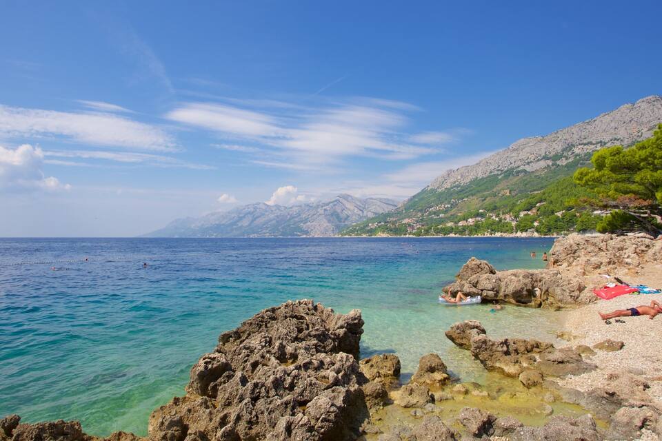 A coastal landscape with clear turquoise waters, rocky shorelines, and a backdrop of mountains.