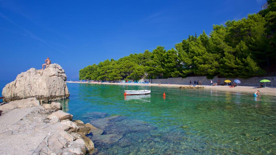 A rocky shoreline with clear turquoise water, a small boat, and a beach with people and umbrellas.
