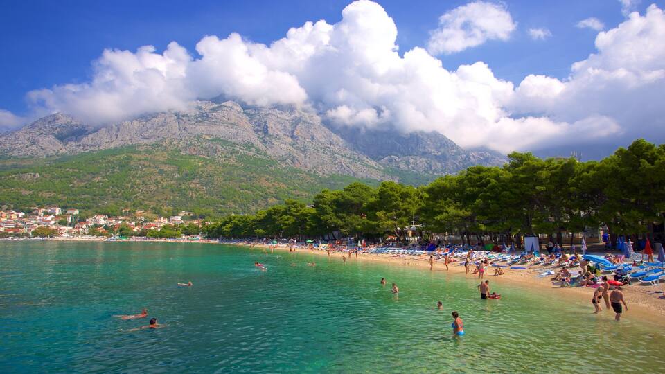 A beach with clear turquoise water, people swimming and sunbathing, and a mountain range in the background.