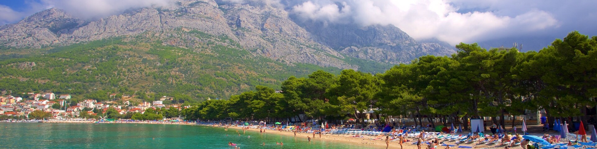 A beach with clear turquoise water, people swimming and sunbathing, and a mountain range in the background.