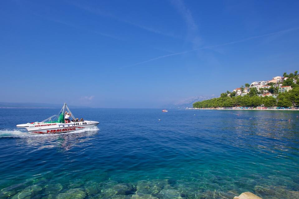 A clear blue sea with a speedboat, rocky shoreline, and buildings on a hillside.