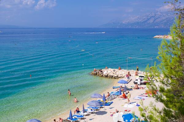 Une plage à l’eau turquoise et limpide, des gens qui se prélassent au soleil, et un voilier à l’horizon.