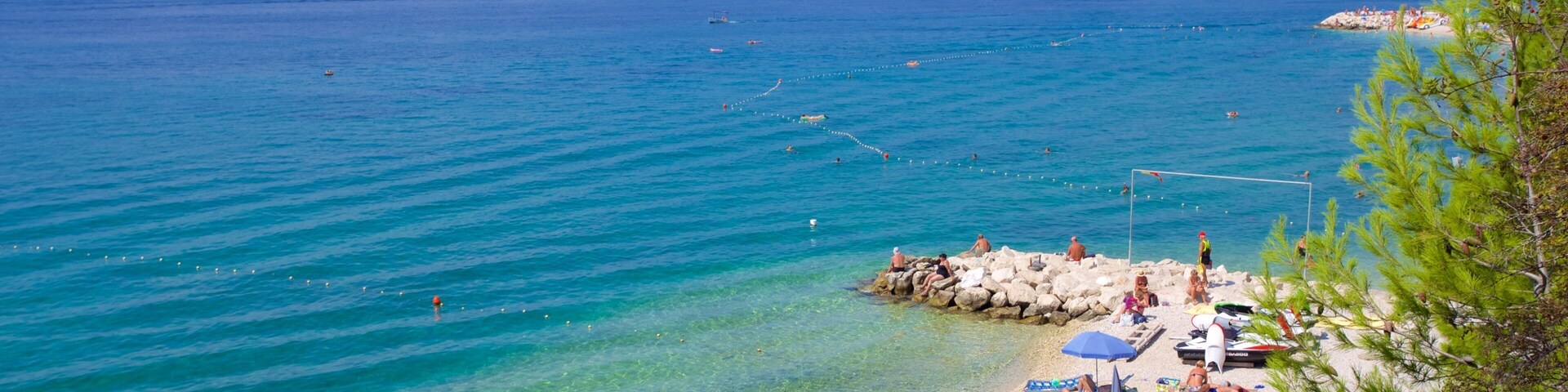 Una playa con agua turquesa cristalina, gente tomando el sol y un velero a lo lejos.