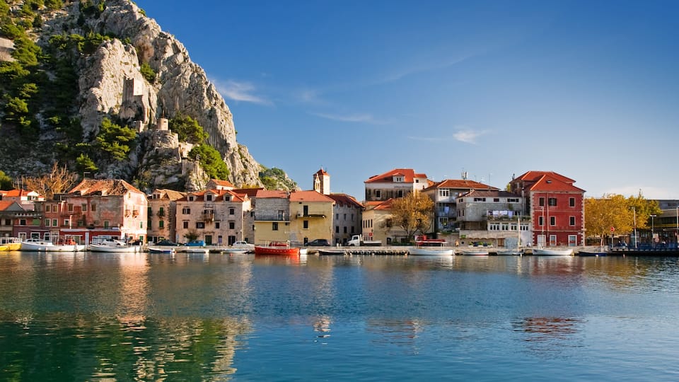 A coastal town with buildings along the water, a rocky cliff, and boats docked at the harbor.