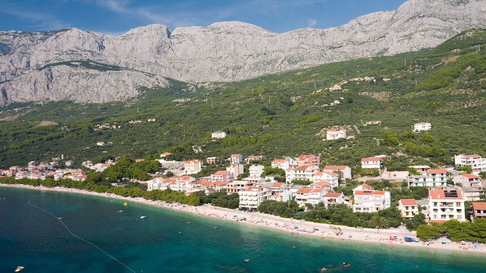 A coastal town with buildings along the beach, mountains in the background, and clear turquoise waters.