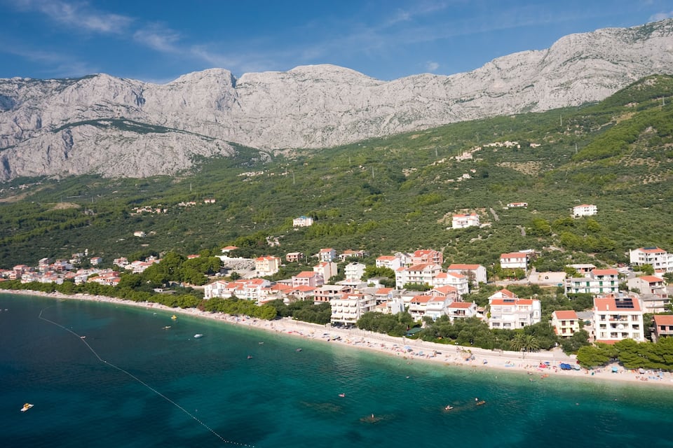 A coastal town with buildings along the beach, mountains in the background, and clear turquoise waters.